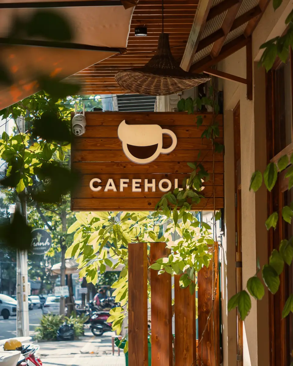 Biển hiệu gỗ Cafeholic giữa giàn cây xanh trước quán